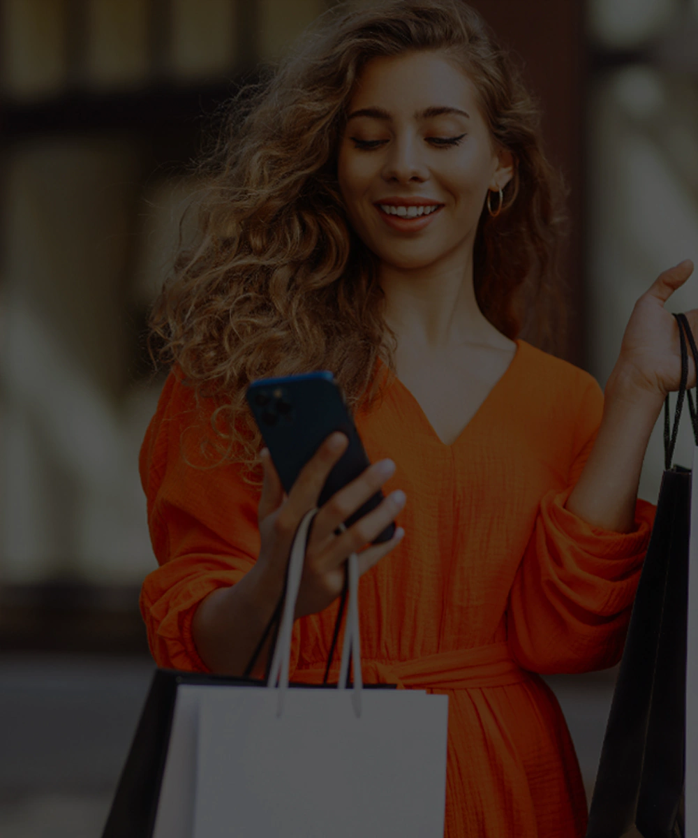 Kobieta idąca w centrum handlowym trzymająca torby zakupowe w jednym ręku i telefon w drugim ręku patrząc na informację z newslettera galerii sudeckiej.