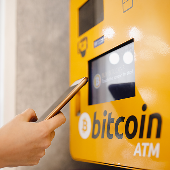 Obraz przedstawia żółty bitomat z napisem bitocin atm, a w kadrze widać również rękę ze smartphonem
