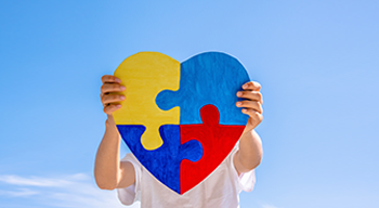 Dłonie chłopca trzymające kolorowe puzzle w kształcie serca na tle błękitnego nieba. Światowy Dzień Świadomości Autyzmu, koncepcja zaburzeń ze spektrum autyzmu.