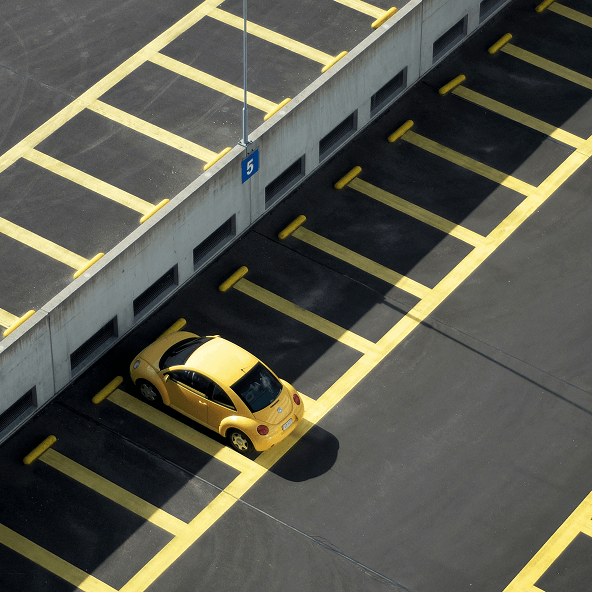 Zdjęcie parkingu z lotu ptaka z wieloma pustymi miejscami i tylko jednym zajętym miejscem, w który stoi zaparkowany żółty samochód.