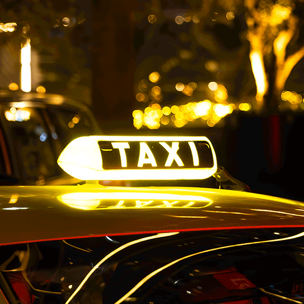 Żółta taksówka z podświetlonym napisem TAXI na dachu. Taksówka stoi zaparkowana nocą w mieście, z rozmazanymi światłami w tle.